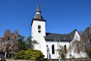 1000 jährige Kirche <p>Weiße Kirche mit Turm vor blauem Himmel, umgeben von blühenden Bäumen und Laterne.</p>