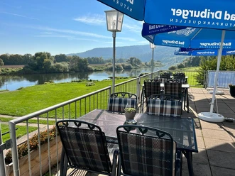Camping am Bootshaus - Terrasse Die Terrasse des Restaurants bietet Tische und Stühle aus Metall, blaue Sonnenschirme und den Blick auf die Weser und Grünflächen.