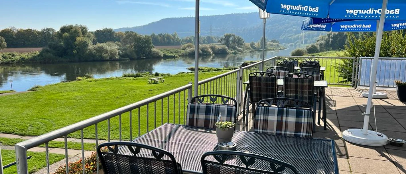 Camping am Bootshaus - Terrasse Die Terrasse des Restaurants bietet Tische und Stühle aus Metall, blaue Sonnenschirme und den Blick auf die Weser und Grünflächen.