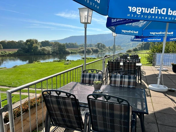 Camping am Bootshaus - Terrasse Die Terrasse des Restaurants bietet Tische und Stühle aus Metall, blaue Sonnenschirme und den Blick auf die Weser und Grünflächen.