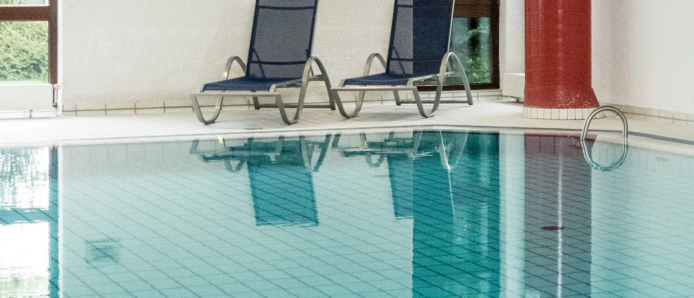 Symbolbild: Schwimmbad Innenpool mit zwei blauen Liegestühlen und großen Fenstern, die natürliches Licht hereinlassen.