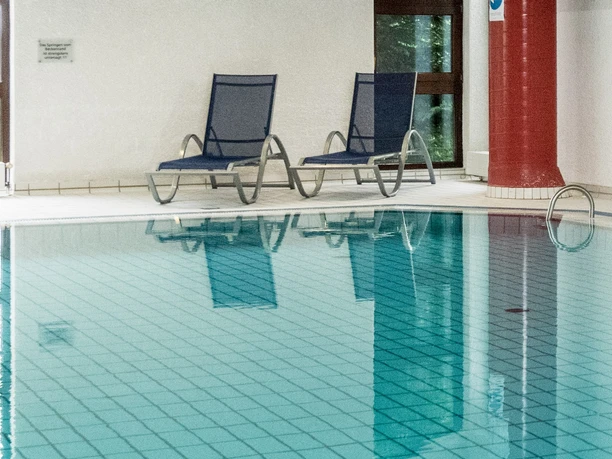 Symbolbild: Schwimmbad Innenpool mit zwei blauen Liegestühlen und großen Fenstern, die natürliches Licht hereinlassen.