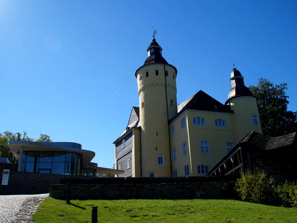 Schloss Homburg Gelbes Schloss mit Türmen neben modernem Gebäude, blauer Himmel im Hintergrund, grüner Rasen davor.