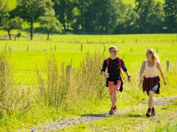 Zwei junge Menschen wandern auf einem sonnigen Feldweg, umgeben von grünem Gras und Bäumen.