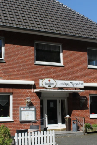 Ein zweistöckiges rotes Backsteingebäude mit weißen Fensterrahmen und einem Schild mit der Aufschrift "Landhaus Wachendorf" über dem Eingang. Vor dem Haus stehen Tische und Stühle auf einer gepflasterten Fläche, umgeben von einem weißen Holzzaun.