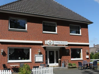 Ein zweistöckiges rotes Backsteingebäude mit weißen Fensterrahmen und einem Schild mit der Aufschrift "Landhaus Wachendorf" über dem Eingang. Vor dem Haus stehen Tische und Stühle auf einer gepflasterten Fläche, umgeben von einem weißen Holzzaun.A two-storey red brick building with white window frames and a sign reading "Landhaus Wachendorf" above the entrance. There are tables and chairs on a paved area in front of the house, surrounded by a white wooden fence.En rød murstensbygning i to etager med hvide vinduesrammer og et skilt med teksten "Landhaus Wachendorf" over indgangen. Der er borde og stole på et asfalteret område foran huset, som er omgivet af et hvidt træhegn.Een rood bakstenen gebouw van twee verdiepingen met witte kozijnen en een bord met "Landhaus Wachendorf" boven de ingang. Er staan tafels en stoelen op een geplaveid terrein voor het huis, omgeven door een wit houten hek.