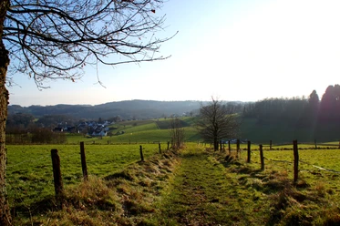 Haferspanienroute durch die Felder Weite Wiesenlandschaft bei Sonnenuntergang, mit Sicht auf ein Dorf und sanfte Hügel im Hintergrund.