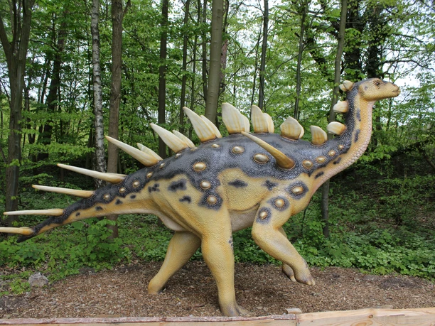 Dino-Park Eine lebensgroße Dinosaurierskulptur steht umgeben von dichtem grünen Laub in einem Waldstück.