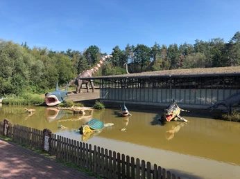 Lebensgroße Dinosauriermodelle am Ufer eines Teiches, umgeben von dichter Vegetation im Dino-Park.