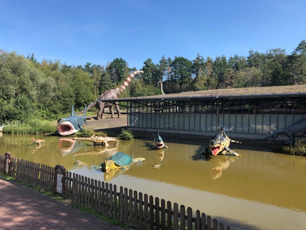 Lebensgroße Dinosauriermodelle am Ufer eines Teiches, umgeben von dichter Vegetation im Dino-Park.
