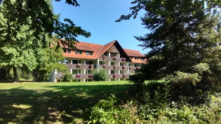 Holsing Vital Mehrstöckiges Gebäude mit roten Ziegeldächern und Balkon, umgeben von Bäumen und blühenden Pflanzen.