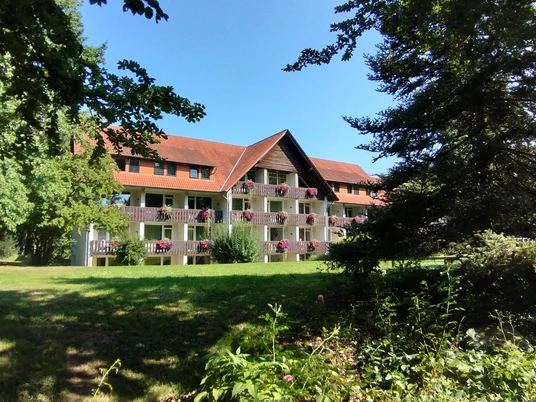 Holsing Vital Mehrstöckiges Gebäude mit roten Ziegeldächern und Balkon, umgeben von Bäumen und blühenden Pflanzen.