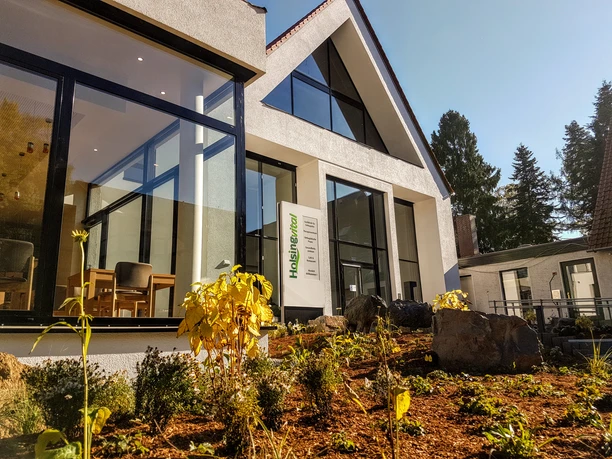 Holsing Vital Modernes Gebäude mit großen Fenstern, gerahmt von Pflanzen, strahlt in hellem Sonnenschein.