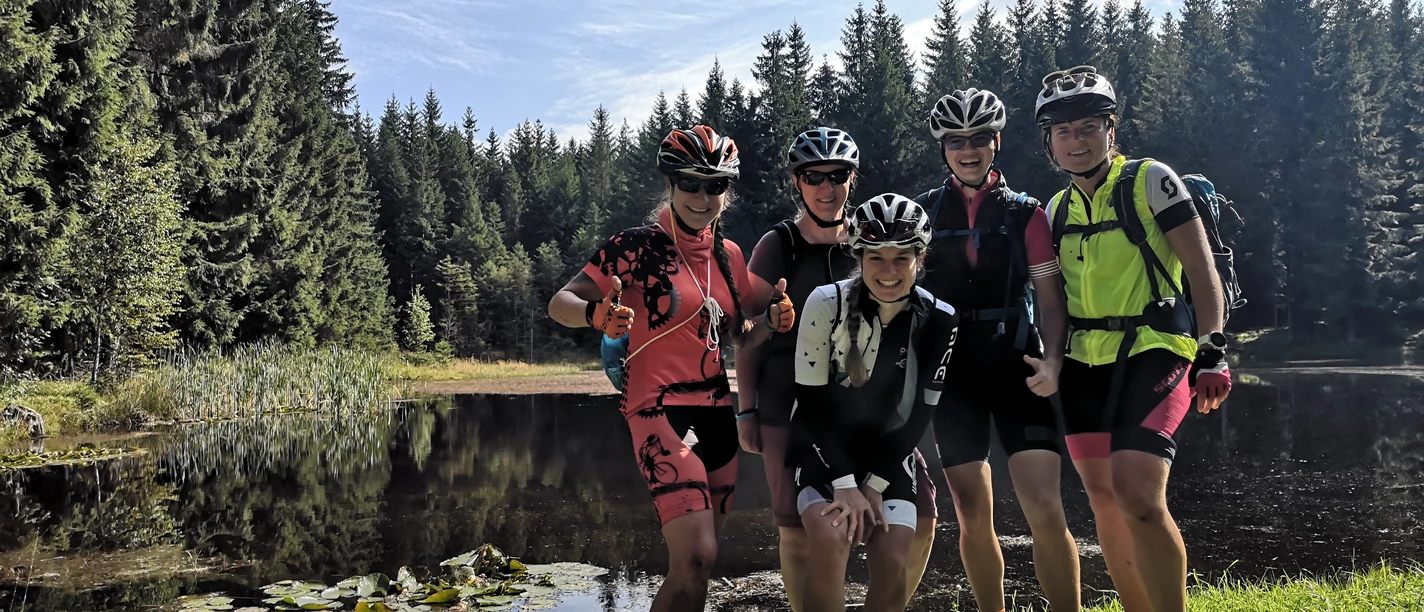IMG_20210905_111557.jpg Fünf Radfahrer in Sportkleidung posieren lächelnd vor einem stillen Waldsee.