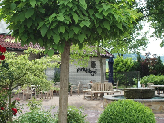 Kurhaus Pivittskrug Sonnige Terrasse des Kurhauses Pivittskrug mit runden Holztischen und üppiger Grünbepflanzung.