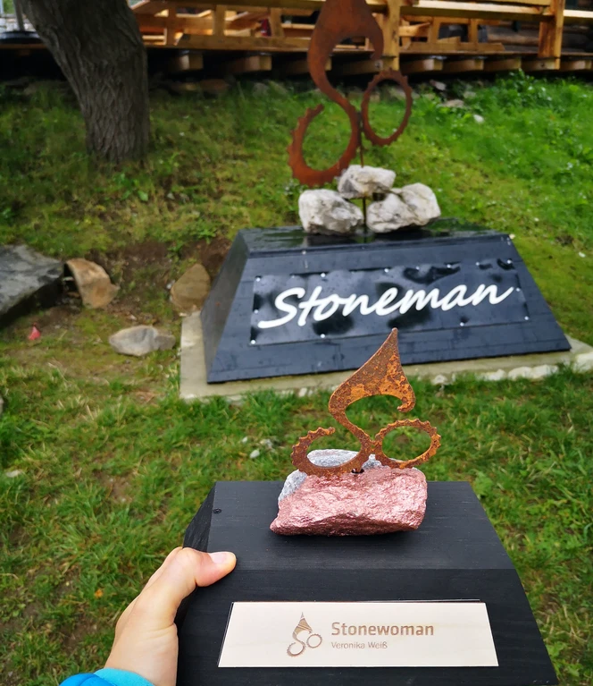 Person holding trophy, statue and "Stonewoman" sign in the green outdoor area.