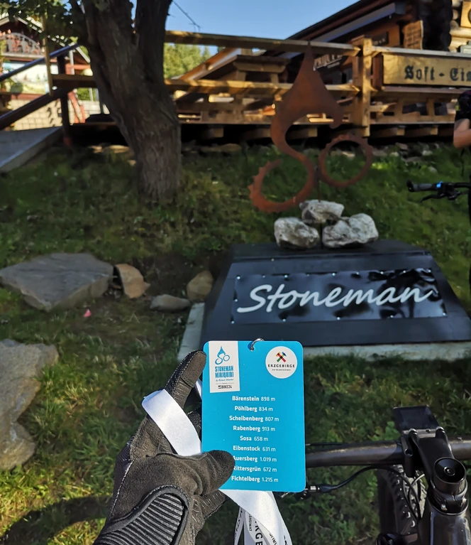 Cyclist holding route badge in front of wooden house with "Stoneman" sign in a green landscape.