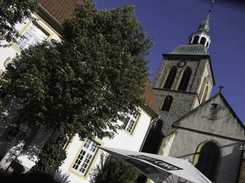 Eine historische Kirche mit Glockenturm und angrenzenden Gebäuden vor einem sonnigen Himmel.