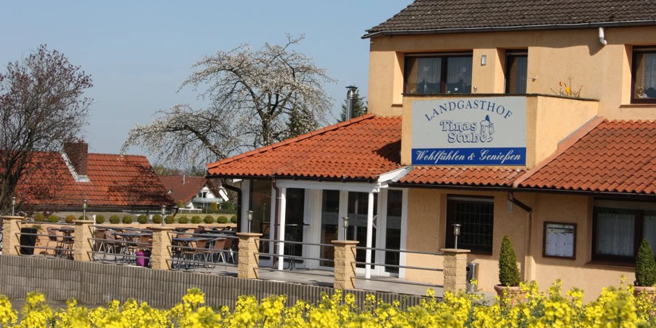 Landgasthof Tinas Stube Zweistöckiges Gebäude mit gelber Fassade und überdachtem Eingang, umgeben von blühendem Rapsfeld.