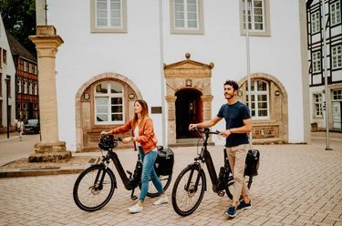 Teutoburger Wald-Brakel-Tourist Info ┬® Teutoburger Wald Tourismus (5).jpg Zwei Personen mit E-Bikes stehen vor einem historischen Gebäude mit Fachwerkelementen.