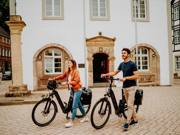 Teutoburger Wald-Brakel-Tourist Info ┬® Teutoburger Wald Tourismus (5).jpg Zwei Personen mit E-Bikes stehen vor einem historischen Gebäude mit Fachwerkelementen.