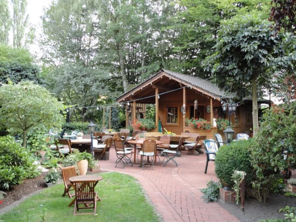 Landhotel und Restaurant zum grünen Kranze Espelkamp.png Ein Landhotel mit Restaurant, umgeben von einem einladenden, grünen Garten mit Holztischen und Stühlen.