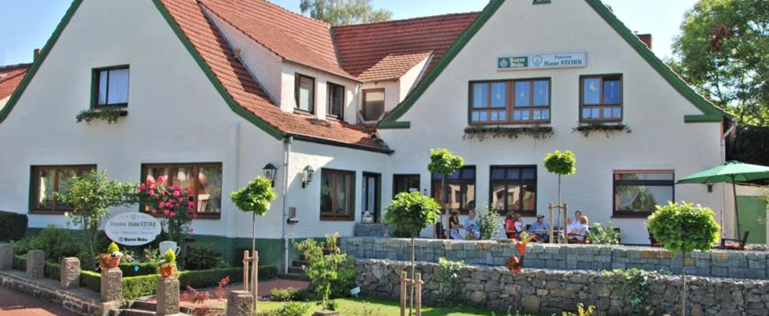 Pension "Haus Stork" Weiße Pension mit roten Dächern und grünem Zierwerk, Terrasse mit Gästen und buntem Garten.