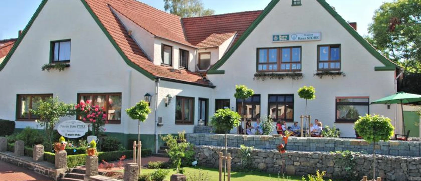 Pension "Haus Stork" Weiße Pension mit roten Dächern und grünem Zierwerk, Terrasse mit Gästen und buntem Garten.