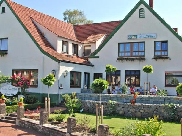 Pension "Haus Stork" Weiße Pension mit roten Dächern und grünem Zierwerk, Terrasse mit Gästen und buntem Garten.