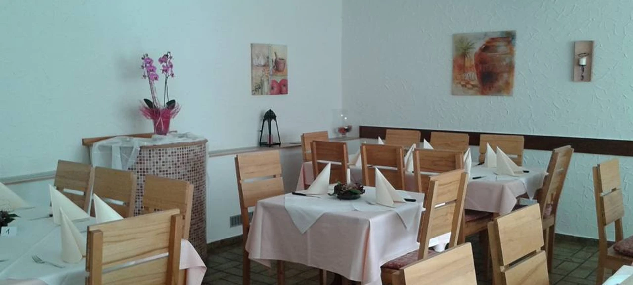 Pizzeria Bella Puglia_PrOldendorf.jpg Heller Gastraum mit Holzmöbeln, weißen Tischdecken und Dekorationen an den Wänden, einladende Atmosphäre.