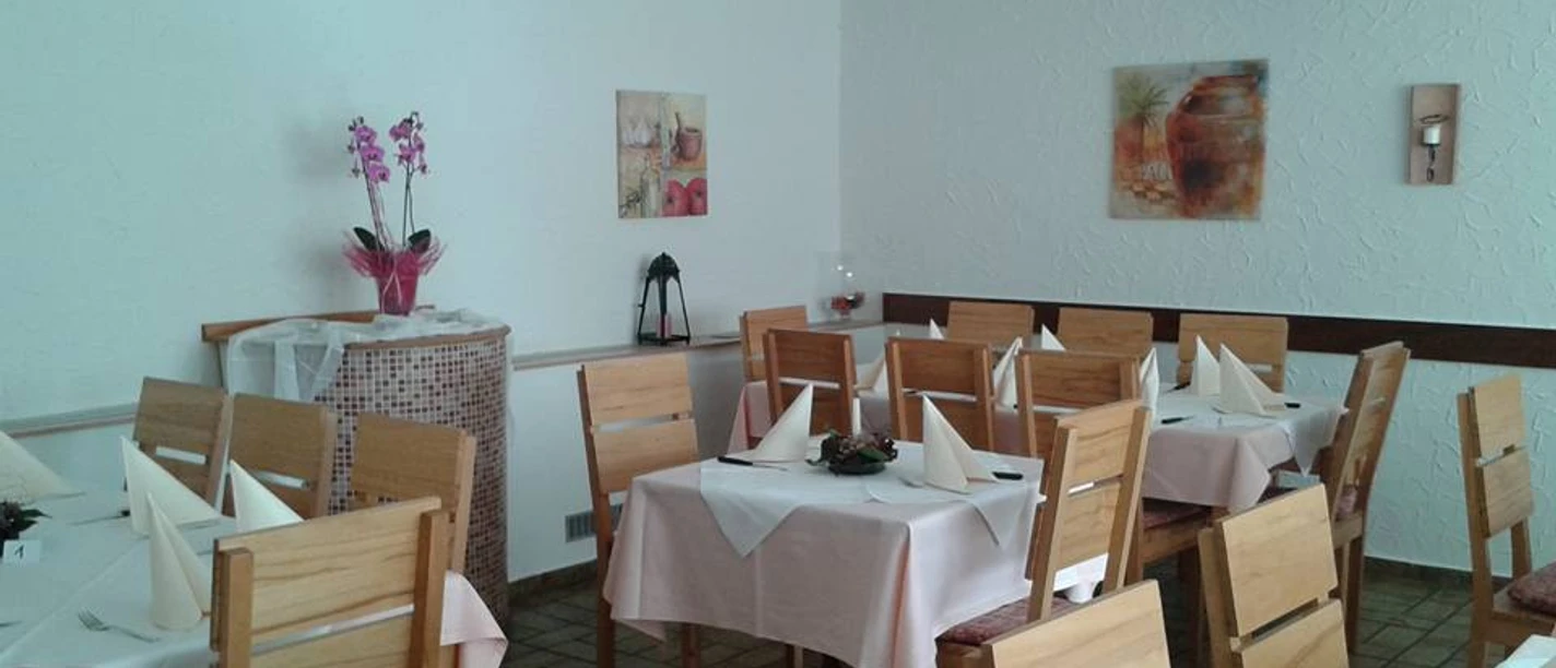 Pizzeria Bella Puglia_PrOldendorf.jpg Heller Gastraum mit Holzmöbeln, weißen Tischdecken und Dekorationen an den Wänden, einladende Atmosphäre.