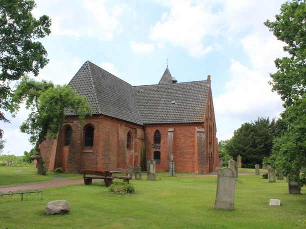 Backsteinkirche mit grauem Ziegeldach in grüner Umgebung, umgeben von Grabsteinen und Bäumen.