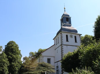 Kirche Blender Historische Backsteinkirche mit schiefergedecktem Turm inmitten grüner Bäume bei strahlendem Himmel.