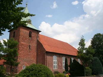 Backsteinkirche mit grünem Turm vor bewölktem Himmel, umgeben von saftigem Grün und Bäumen.