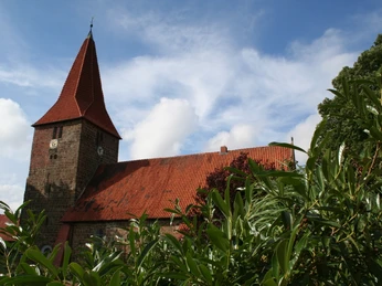 St. Bartholomäus Kirche Balge Rote Ziegeldachkirche mit markantem Turm, umgeben von grünem Laub unter einem blauen Himmel.