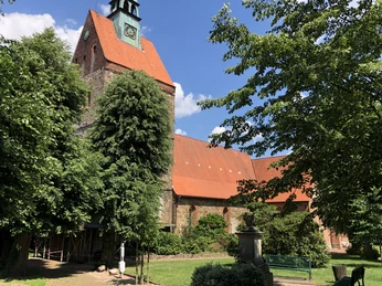 A historic church with a red-tiled roof and tower, surrounded by trees, under a blue sky.