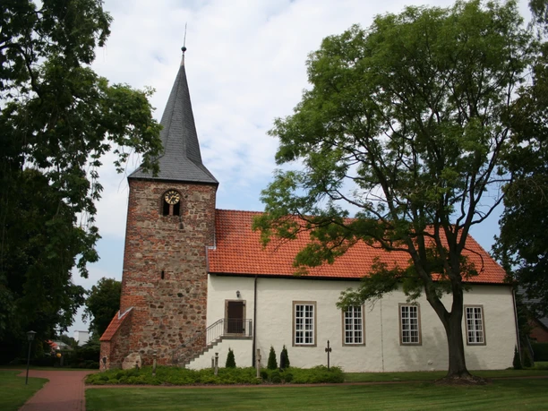 Kirche Warmsen Eine historische Kirche mit rotem Ziegeldach, markantem Turm und umgeben von grünen Bäumen.