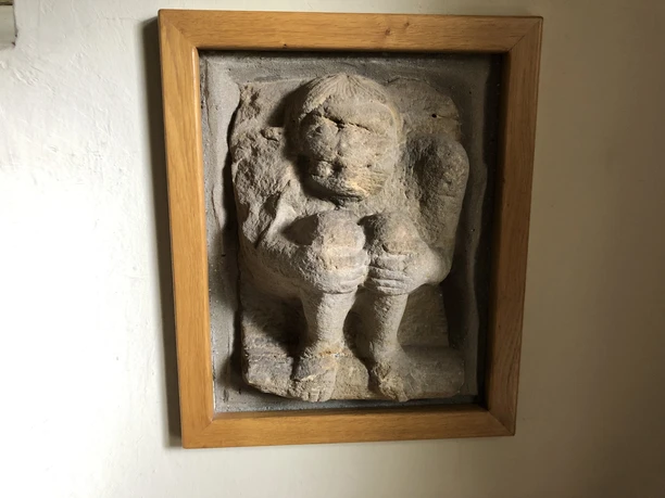 Hockender Mönch Reliëf van een hurkende monnik in een houten frame, gemaakt van zandkleurige steen, op een muur.