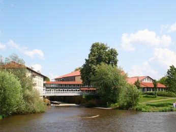 Hotel mit rotem Dach an Flussufer, umgeben von grünen Bäumen, blauer Himmel im Hintergrund.