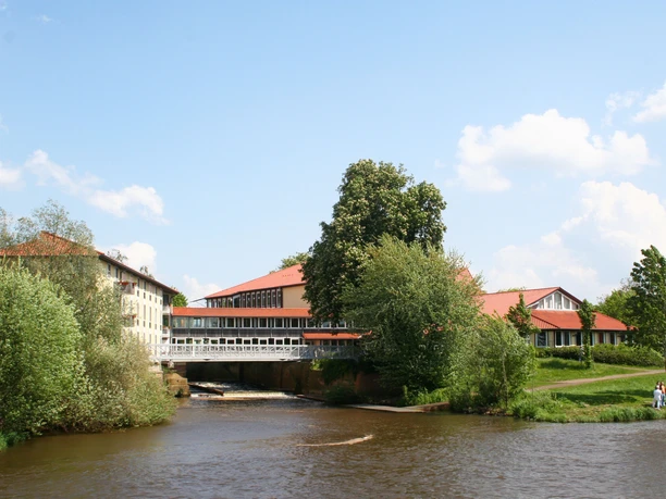 Hotel with red roof on the riverbank, surrounded by green trees, blue sky in the background.