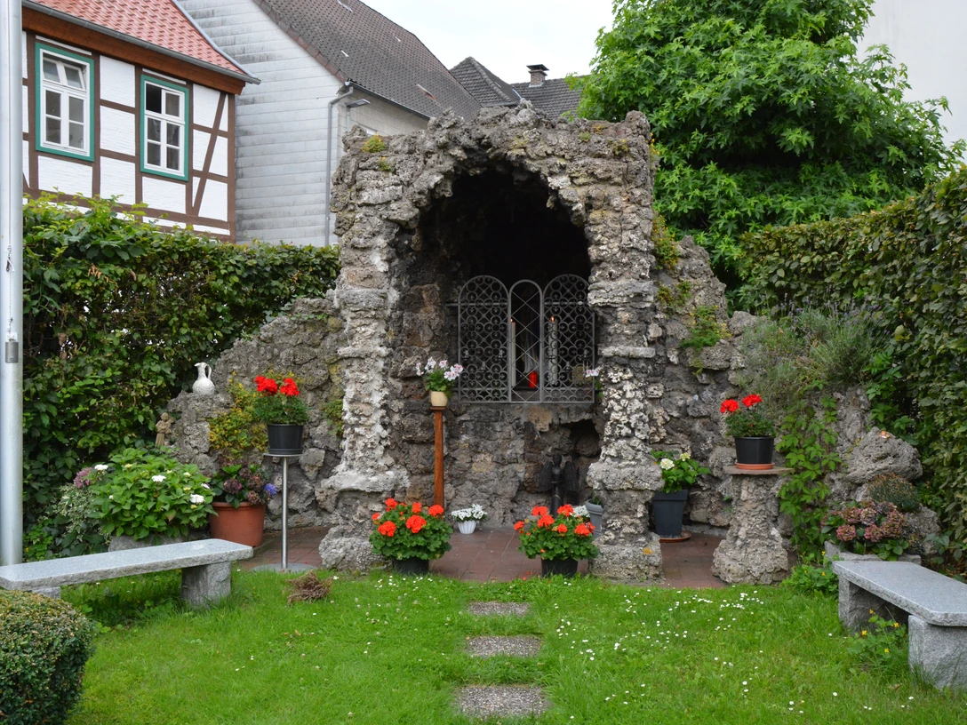 Eine mit Blumen geschmückte Grotte aus Naturstein, umgeben von gepflegtem Grün und zwei Steinbänken.