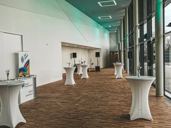 Moderner Empfangsraum mit hohen Glasfenstern, eleganten Stehtischen und einer Bühne im Hintergrund.