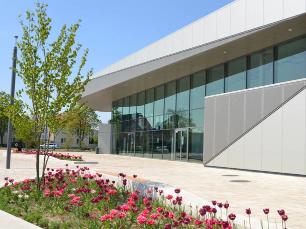 Modernes Gebäude mit großer Glasfront, umgeben von pinken Blumenbeeten und jungen Bäumen.