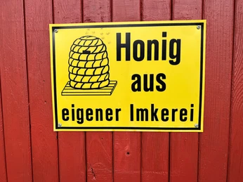 Honigschild Gelbes Schild auf roter Holzwand mit schwarzer Aufschrift: "Honig aus eigener Imkerei" und Bienenmotiv.