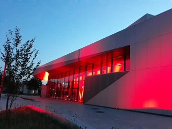 Stadthalle Night of light Modernes Gebäude bei Abenddämmerung mit rot beleuchteter Glasfassade, umgeben von Pflanzen.