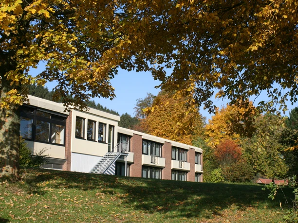 Moderne Therme mit großen Fenstern, umgeben von buntem Herbstlaub und einem klaren, blauen Himmel.