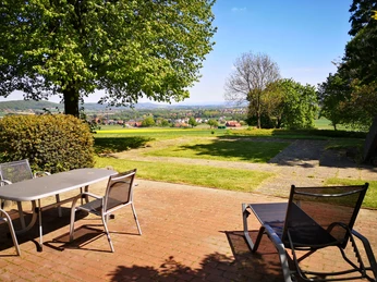 Wiehen Therme Terrasse mit Tisch und Stühlen im Vordergrund, bietet Blick auf eine grüne Landschaft und entfernte Hügel.