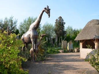 Metallskulptur einer Giraffe mit umliegender Vegetation und einem Strohdachpavillon im Hintergrund.