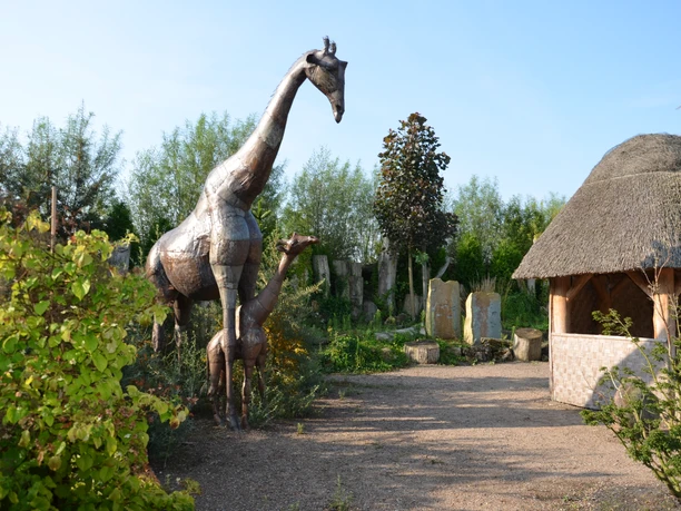 Rila erleben - Afrika Metallskulptur einer Giraffe mit umliegender Vegetation und einem Strohdachpavillon im Hintergrund.