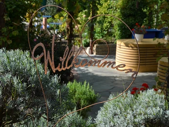 Rila erleben Ein herzförmiges Metallschild mit "Willkommen", umgeben von grünen Pflanzen in einem Garten.